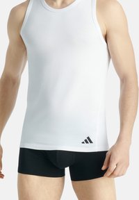 Bílý bezrukávový top s jemným leskem a černým logem Adidas na spodní části. Modelka má na sobě černé těsné kraťasy.