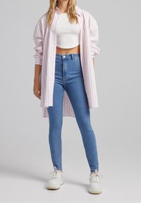 Kvinna som bär en ljusrosa och vit randig lång skjorta, vit crop top, blå tajta jeans och vita sneakers som står mot en enkel bakgrund.