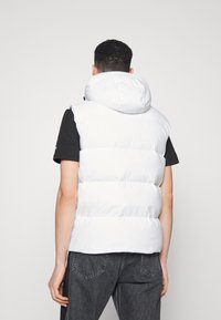 Vit pufferväst med huva, med en kviltad design, slät textur och ärmlös stil. Bärs över en svart t-shirt och grå jeans.