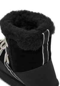 Fekete csizma, amely textúrált, bolyhos gallérral, velúrbőr felsőrésszel és kontrasztos fehér cipőfűzőkkel rendelkezik. A hátsó részen bordás szövet díszítéssel van ellátva.