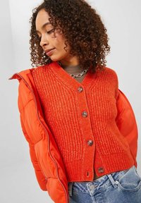 Cardigan arancione a coste con scollo a V, cinque bottoni e tessuto testurizzato, abbinato a un giubbotto imbottito arancione brillante e jeans di denim.