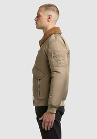 Ung mand i profil iført en beige bomberjakke med brun sherpa-krave og sorte bukser, stående op ad en ensfarvet lys baggrund.