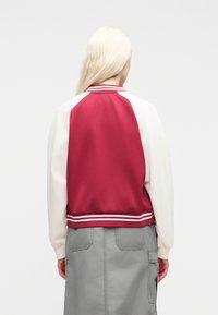 Άτομο με μακριά ξανθά μαλλιά που φοράει κόκκινο και άσπρο πανωφόρι varsity και γκρι φούστα, εμφανιζόμενο από πίσω σε απλό φόντο.