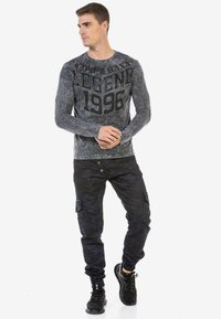Graues Langarmshirt mit dem Aufdruck „LEGEND 1996“, kombiniert mit dunklen camouflage Cargo-Hosen und schwarzen Sneakers. Lässiges, figurbetontes Design.