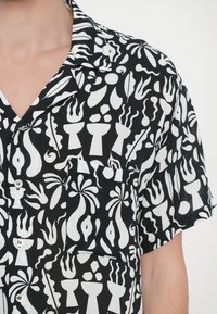 Μαύρο κοντομάνικο πουκάμισο με λευκό αφηρημένο σχέδιο, διαθέτει τσέπη στο στήθος και κλείσιμο με κουμπί, κατασκευασμένο από ελαφρύ ύφασμα.