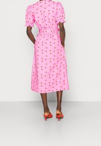 Robe midi rose avec des manches courtes ballon, taille cintrée et motif fraise. Portée avec des chaussures à talons rouges. La vue arrière met en valeur le nœud à la taille.