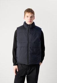 Gilet puff d'un merlo scuro con colletto alto, chiusura frontale con zip e due tasche orizzontali sul petto, caratterizzato da una texture liscia e un materiale leggero.