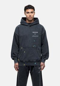 Zwarte oversized hoodie gemaakt van een katoenmix, voorzien van een voorzak, verfsplatters en gedrukt tekst. Zachte textuur met een casual ontwerp.