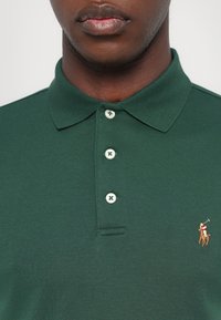 Mørkegrønn polo skjorte laget av glatt bomull med kragehals, med tre hvite knapper og en liten brodert logo på venstre bryst.