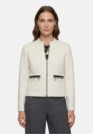 Femme portant une veste zippée blanche texturée avec des poches zippées noires sur un haut à motifs et un pantalon sombre, debout devant un fond uni.