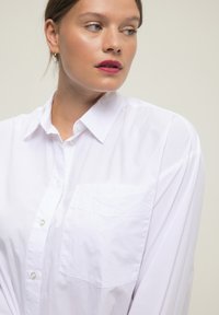 Camisa blanca de botones, hecha de una tela ligera, con cuello, mangas largas y un bolsillo en el pecho. Textura suave.