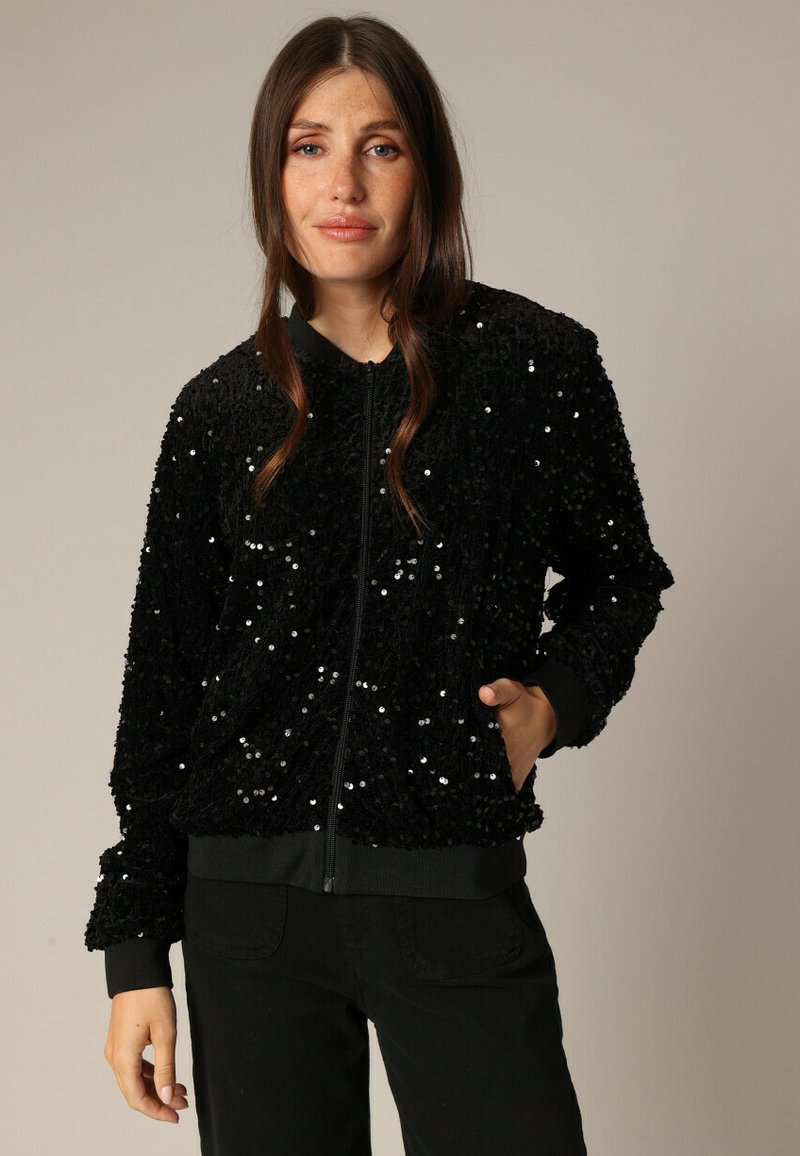 Veste bomber noire en tissu texturé, recouverte de sequins brillants. Elle présente une fermeture éclair à l'avant, des poignets côtelés et une coupe décontractée.