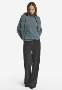 Blaugrüner geschichteter Reißverschluss-Weste über einem passenden Rollkragenpullover, kombiniert mit hochtaillierten schwarzen weiten Jeans. Glatte Textur und minimalistisches Design.