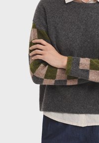 Grijze wollen trui met een relaxte pasvorm, met mouwen in een groen en roze geruit patroon. Gelaagd over een witte blouse met kraag.