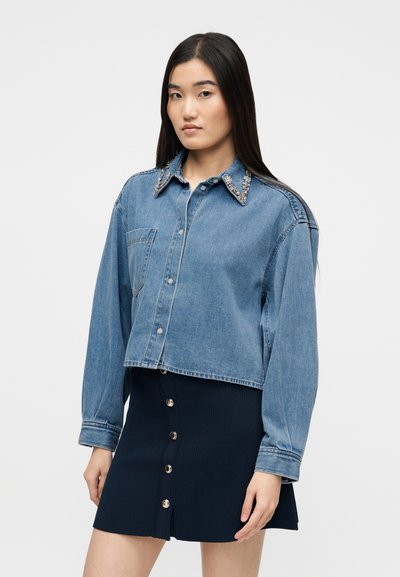 Γυναίκα που φοράει ένα κομμένο μπλε τζιν τζάκετ με στολισμένο γιακά και μια μαύρη μίνι φούστα με κουμπιά, στέκεται μπροστά σε λευκό φόντο.