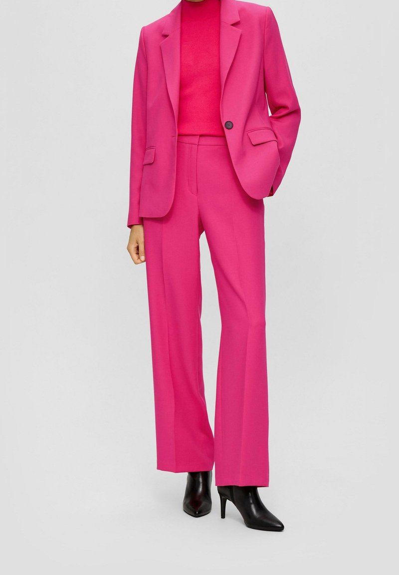 Femme portant un tailleur rose vif composé d'un blazer et d'un pantalon large, associé à des bottines noires à talons aiguilles pointues.