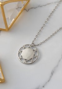 Sølv halskæde med en cirkulær vedhæng med stjernedesign og en hvid, glat midte, placeret på en marmoroverflade.