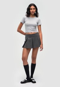 T-shirt grigio corto abbinato a una gonna a pieghe grigio scuro. Il modello indossa calze nere al ginocchio e scarpe di vernice nera. Sfondo semplice.