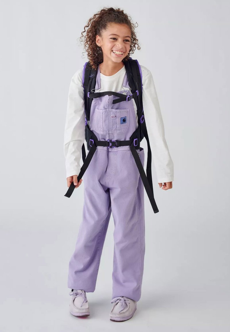 Smiling child wearing lavender overalls and white long-sleeve shirt, holding black backpack straps, standing against a plain background.