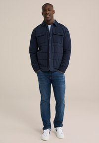 Tengerészkék steppelt, gombos kabát két mellzsebbel, kék farmerrel és fehér tornacipővel párosítva. Sima anyag, strukturált kialakítás.