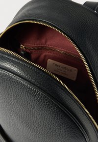 Mochila de cuero negro con superficie texturizada y cremallera dorada. El interior cuenta con un forro granate y una pequeña etiqueta de cuero con la inscripción "Coccinelle".