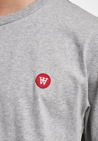 Szary bawełnowy t-shirt z prążkowanym okrągłym dekoltem i małą czerwoną okrągłą naszywką z białą literą "W" haftowaną na piersi.
