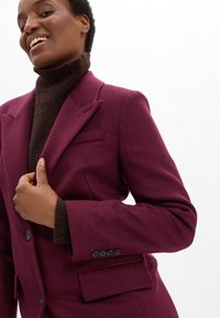 Een lachende persoon draagt een bordeauxkleurige blazer over een donkerbruine coltrui, terwijl hij met één hand de revers van de blazer vasthoudt, tegen een witte achtergrond.