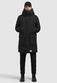 Sort polstret parka med høj krave, justerbar hætte, to frontlommer og lynlåslommer. Bæres sammen med sorte bukser og støvler.