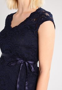 Marinblå klänning i spets med korta ärmar och v-ringning. Har ett texturerat blommönster och ett satinband knutet i midjan.