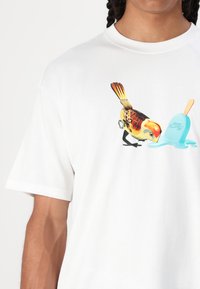 Hvid t-shirt i bomuld med en farverig fugl illustation og en blå is med en orange stik, centreret på forsiden.