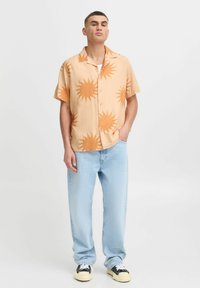 Kurzärmliges, hellorangefarbenes Hemd mit Sonnenmuster; kombiniert mit hellblauen Jeans und schwarzen Sneakers. Lässiger Schnitt und entspanntes Design.
