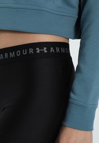 Czarne legginsy dopasowane z teksturowanej tkaniny i szerokim pasem w talii z szarym napisem "ARMOUR", w zestawie z jasno niebieską krótką bluzą.