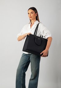 Black tote bag with textured material, two handles, and a structured shape. Featuring minimalistic design and gold logo accent.