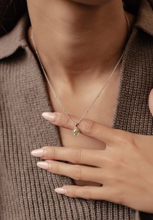 Silver necklace with a pear-shaped green gemstone pendant, detailed with small clear stones, worn against a textured brown sweater.