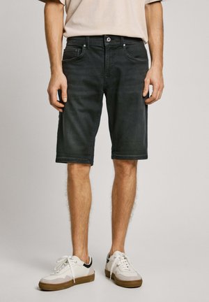 Homme portant un short en denim noir, une chemise beige et des baskets blanches à semelles en caoutchouc, debout devant un fond gris clair uni.