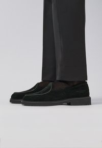 Black suede slip-on shoes with a smooth texture, featuring a contrasting trim, paired with tailored black trousers.