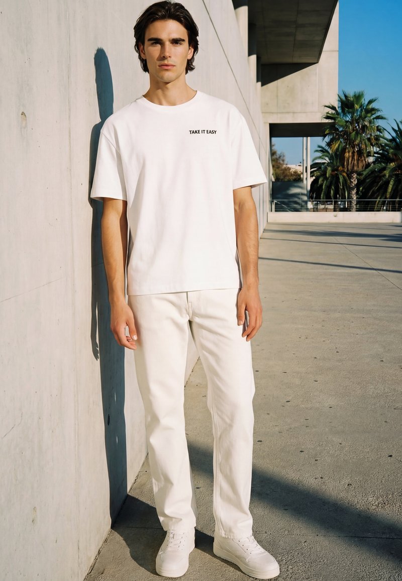 Jeune homme en t-shirt blanc avec le texte "TAKE IT EASY", pantalon blanc et baskets blanches, debout contre un mur en béton à l'extérieur au soleil.