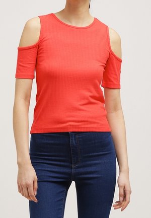 Vrouw met een felrood off-shoulder top met korte mouwen en donkerblauwe jeans, staand tegen een effen lichte achtergrond.
