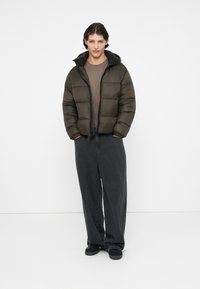 Chaqueta acolchada en verde oliva con forro negro, diseño acolchado, puños elásticos y cremallera. Combinada con pantalones de pierna ancha gris oscuro.