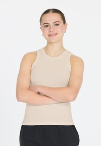 Beige gerippter Tanktop mit runder Ausschnittform und ärmellosem Design, kombiniert mit schwarzen Hosen. Der Stoff wirkt weich und dehnbar.
