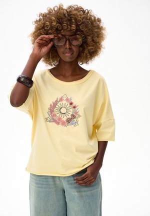 Kobieta z kręconymi włosami, w dużych okularach przeciwsłonecznych, żółtej koszuli z motywem słońca i kwiatów, w niebieskich dżinsach oraz czarnej bransoletce, poprawiająca okulary.