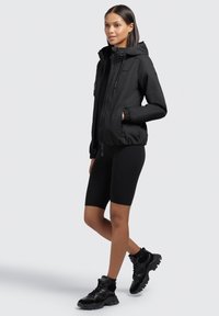 Veste noire légère zippée avec capuche, poignets élastiques et poches latérales, associée à un short noir et des baskets noires imposantes.