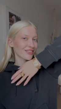 Jeune femme aux longs cheveux blonds souriante, portant un sweat à capuche noir, une montre en or et une bague en or, posant en intérieur avec des œuvres encadrées en arrière-plan.