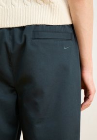 Pantaloni negri Nike cu o textură netedă, având un buzunar în spate și un logo subtil în culoarea asortată. Pulover bej tricotat vizibil deasupra.
