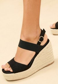 Sandali wedge neri con tomaia in tela testurizzata, cinturino elasticizzato alla caviglia e un plateau in juta intrecciata. Design open-toe con una finitura leggermente lucida.
