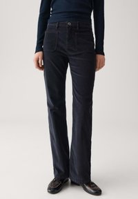 Persoon in donkere hoge corduroy broek, zwarte schoenen en een donker langwerpig ribbelshirt, staande tegen een effen achtergrond.