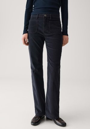 Persoon in donkere hoge corduroy broek, zwarte schoenen en een donker langwerpig ribbelshirt, staande tegen een effen achtergrond.