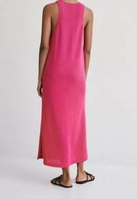 Robe longue rose en maille, sans manches avec une fente latérale. Présente une coupe décontractée et un tissu texturé. Portée avec des sandales plates noires.