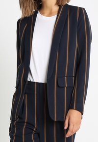 Granatowa marynarka w brązowe pionowe paski, dopasowany krój, zapięcie na jeden guzik i dwie przednie kieszenie, noszona na białym t-shircie.