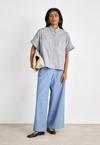 Camicia a righe, a forma di scatola, con due tasche sul petto, abbinata a pantaloni larghi di un blu chiaro e scarpe nere basse. Tiene una borsa a tracolla beige.